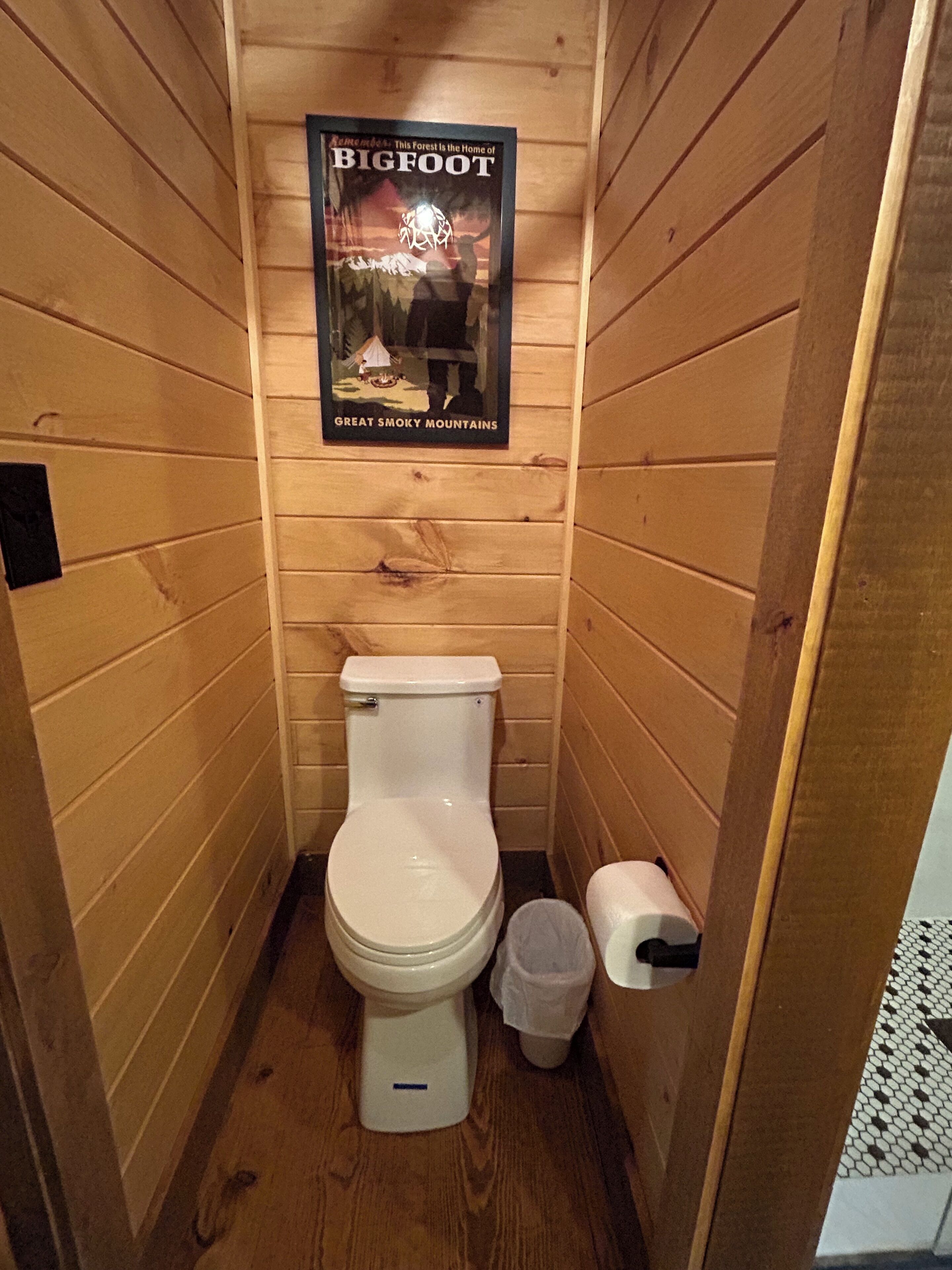 A small wooden cabin-style bathroom with a toilet, a trash bin, and a poster on the wall.