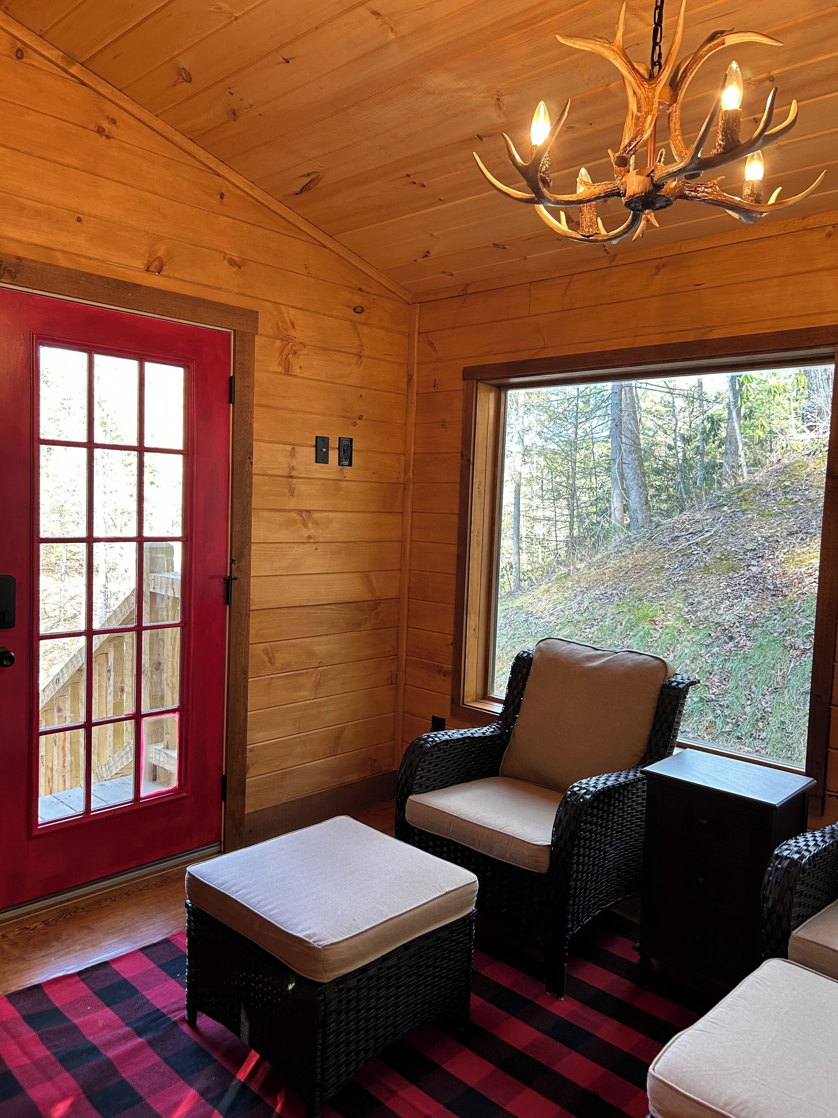 Ein gemütliches Zimmer mit roter Tür, Korbmöbeln, einem karierten Boden und einem großen Fenster mit Blick auf Bäume.