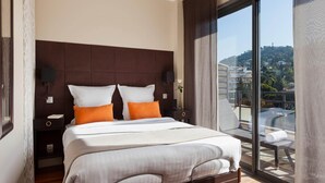 Un dormitorio de hotel moderno con una cama grande, dos mesitas de noche, vistas de la ciudad a través de amplios ventanales y un balcón con mobiliario exterior.