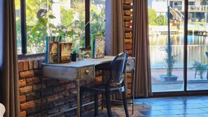 A room with a brick wall, a desk with a chair, a window with curtains, and a view of a pool and outdoor area. - Waterfront Property With A Jetty (Sussex Inlet)
