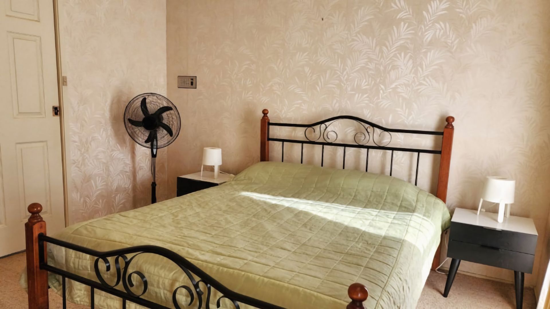 Una camera da letto con un letto, un comodino, una lampada da comodino, una ventola a parete e una porta.