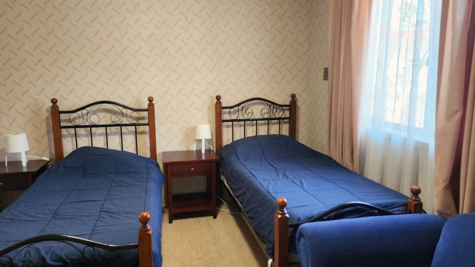 A room with two single beds, each with a wooden headboard and a blue mattress. There is a nightstand with a lamp between the beds.