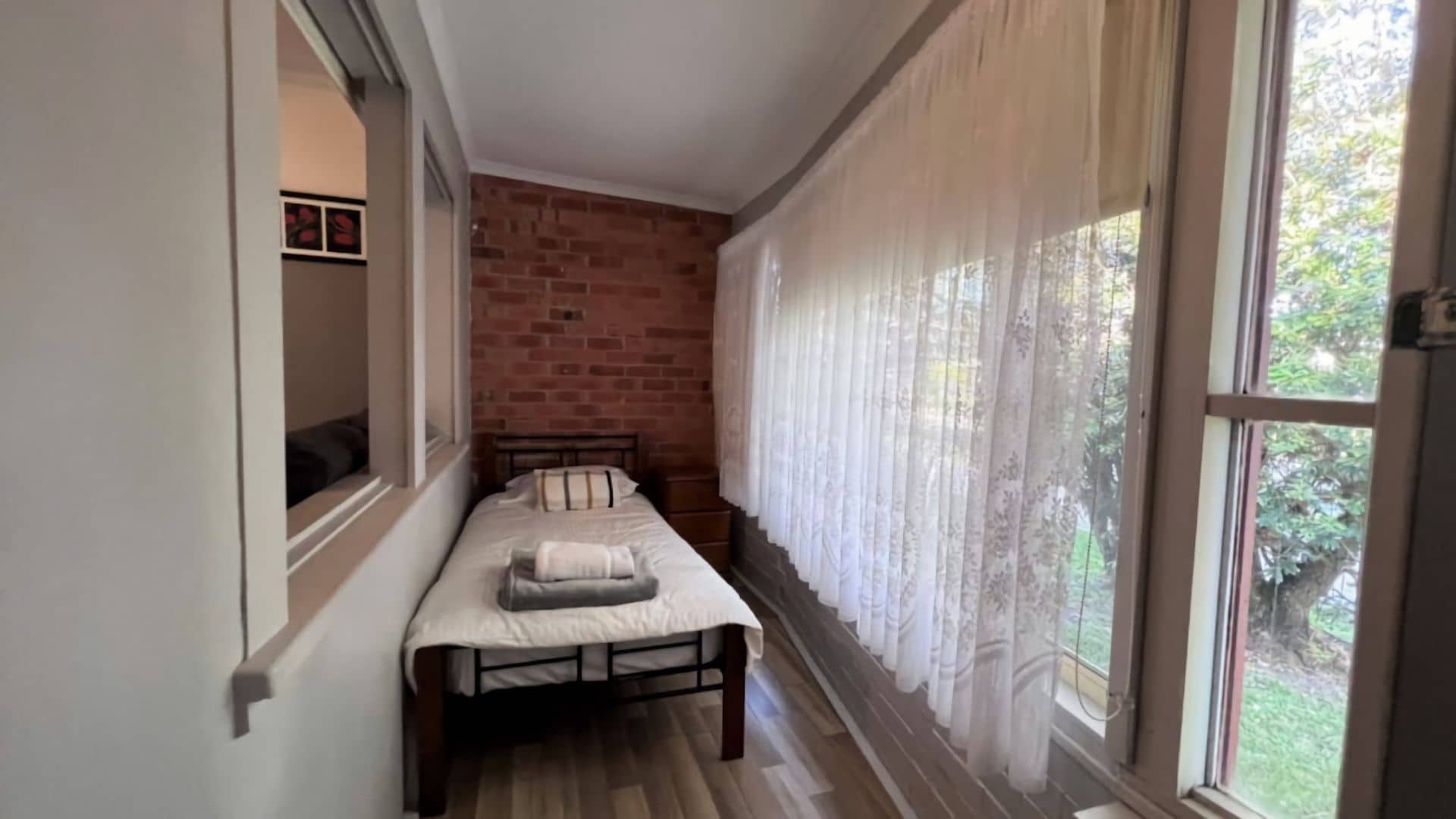 Ein schmales Zimmer mit einem Bett, einer Ziegelwand und einem Fenster mit Vorhängen.