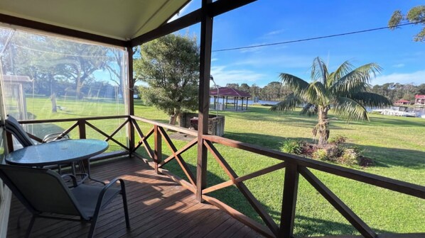 A covered outdoor area with a table and chairs, a wooden deck, and a view of a grassy field with trees and a body of water. - Cottage 3 ~ Family cottage at Sussex House Holiday Park (Sussex Inlet)
