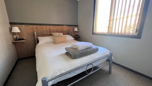 Una cama bien tendida con almohadas y una manta doblada, una mesita de noche con una lámpara y una ventana con persianas.