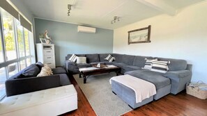 Ein Wohnzimmer mit einem Ecksofa, einem weißen Hocker und einem Couchtisch. Ein Fenster lässt natürliches Licht herein, und an der Wand hängt ein Gemälde.