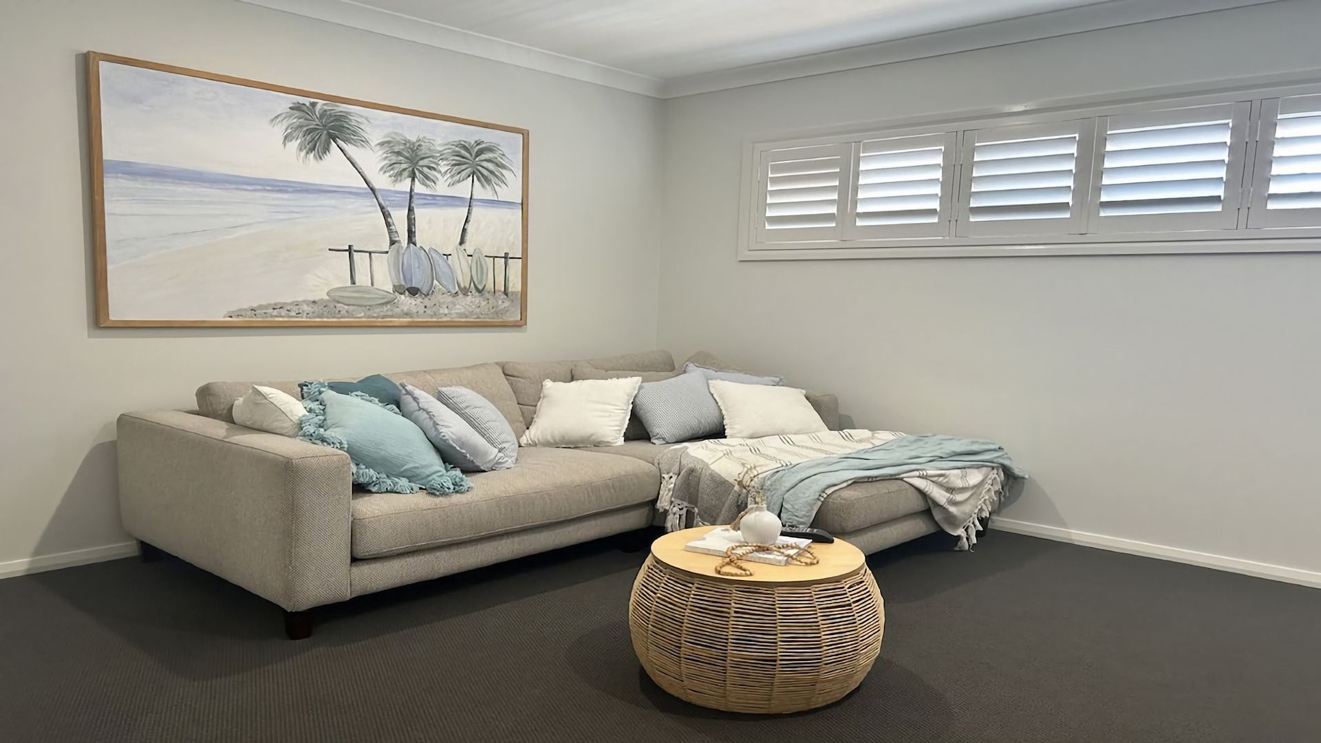 A living room with a beige sectional sofa, a round wicker coffee table, and a beach-themed painting on the wall.