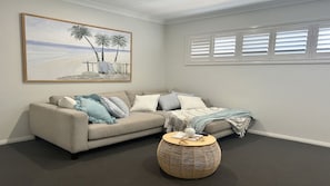 Ein Wohnzimmer mit einem beigefarbenen Ecksofa, einem runden Kaffeetisch aus Rattan und einem Wandbild mit Strandmotiv.