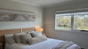 Ein Schlafzimmer mit Bett, Kissen, einer Nachttischlampe und einem Fenster mit Blick auf Bäume.