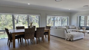 A spacious living area with a dining table and chairs, a sofa, and large windows offering a view of trees.