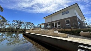 A modern two-story house with a large window, a stone pathway, and a body of water in the foreground. - Sandy Quays on the water (Sussex Inlet)