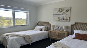 A bedroom with two single beds, a window with a view, a wooden headboard, a bedside table with lamps, and a painting on the wall. - Sandy Quays on the water (Sussex Inlet)