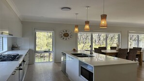 A modern kitchen with a central island, a dining area with wicker furniture, and a large window with a view of trees. - Sandy Quays on the water (Sussex Inlet)