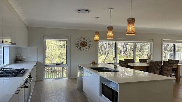 A modern kitchen with a central island, a dining area with wicker furniture, and a large window with a view of trees.