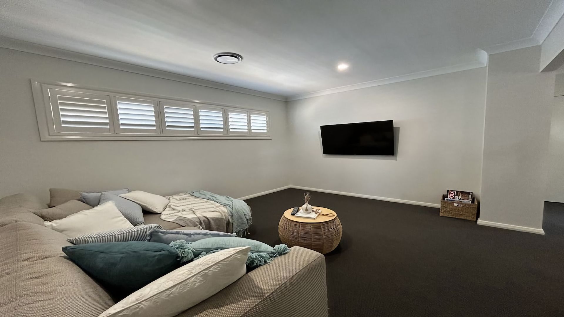 A living room with a sofa, pillows, a round coffee table, a flat-screen TV mounted on the wall, and a window with blinds.