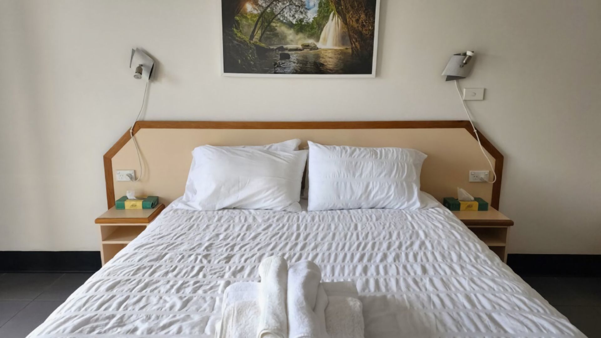 A neatly made bed with white linens and pillows, a wooden headboard, and a painting of a waterfall on the wall.