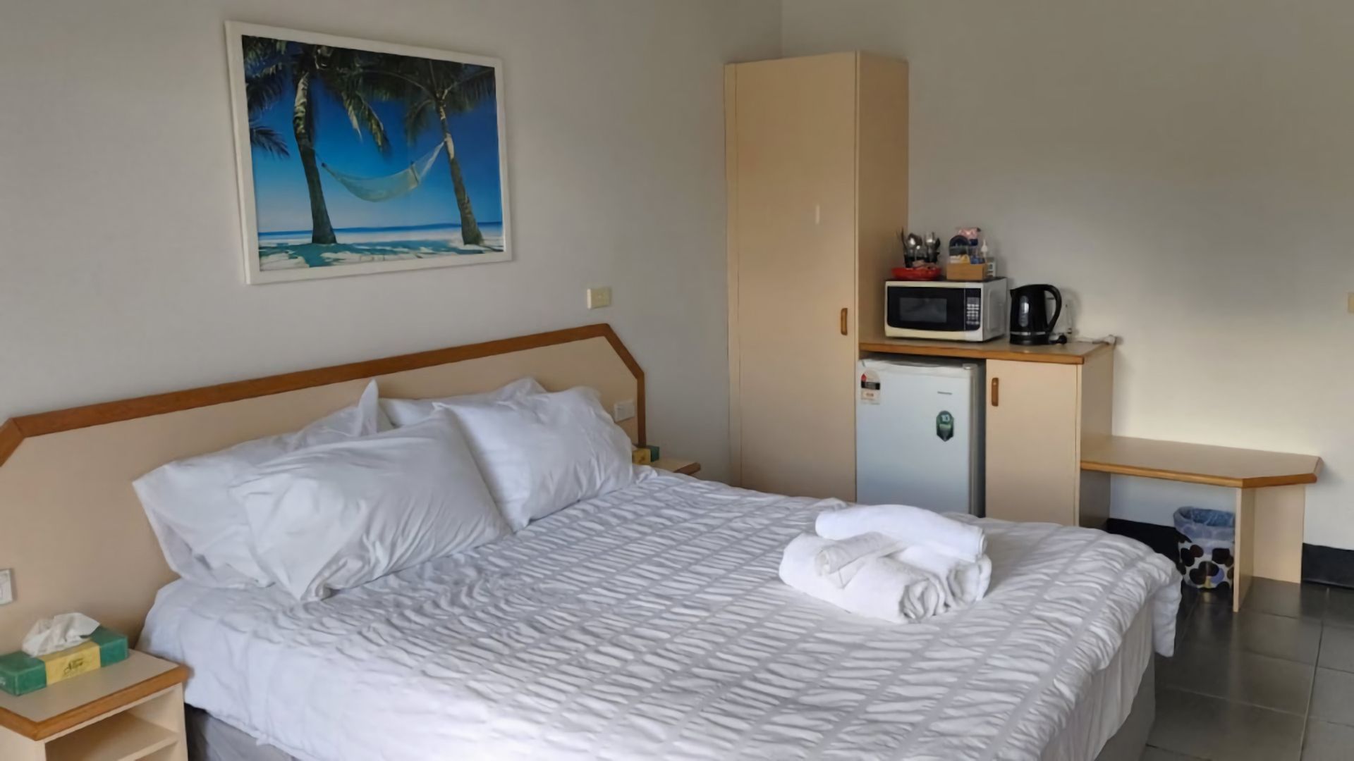 A hotel room with a bed, a painting of a beach, a kitchenette, and a small refrigerator.