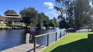 A riverside walkway with a wooden deck, a grassy area, and a building in the background.