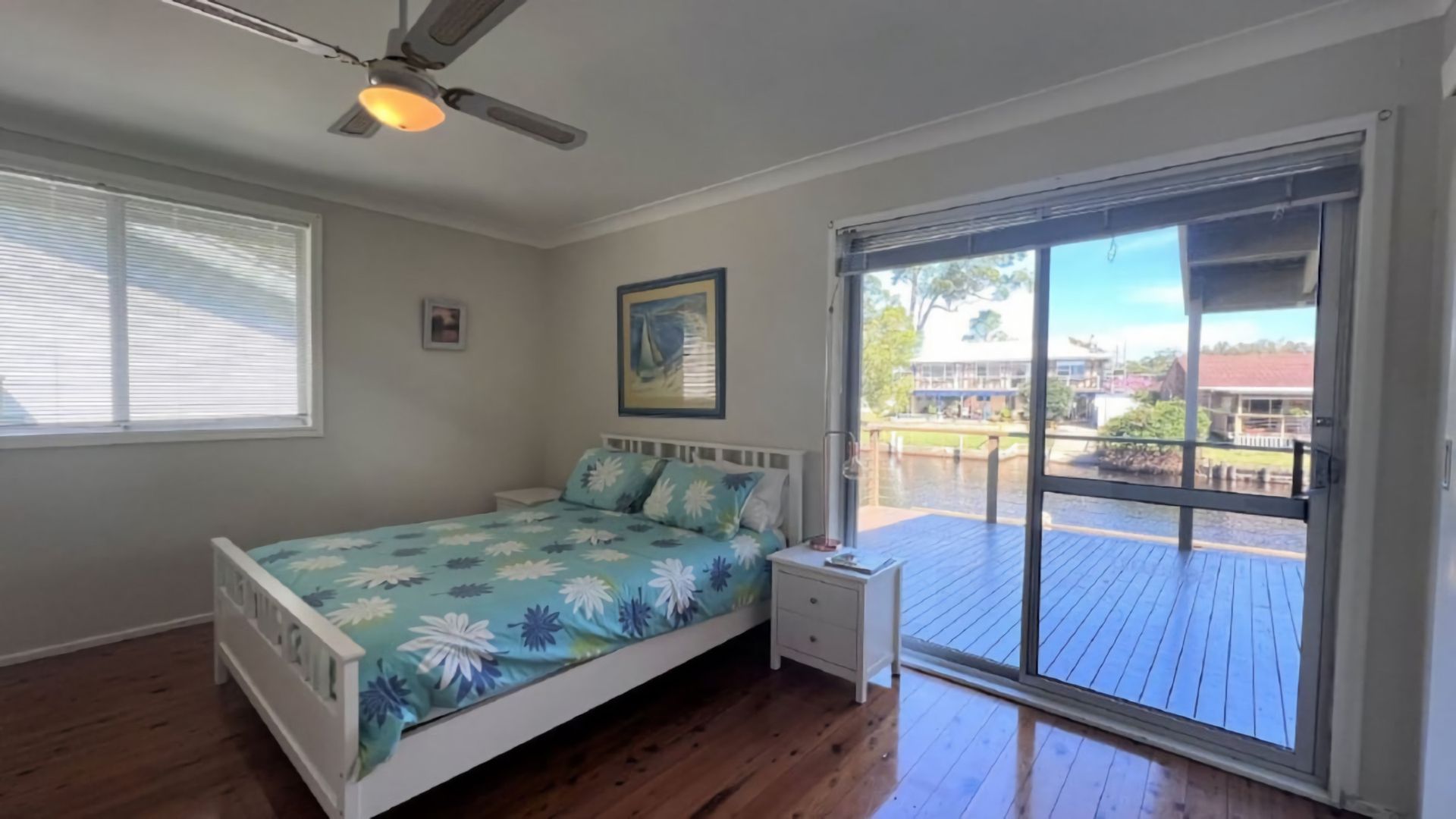 A bedroom with a bed, a nightstand, a ceiling fan, a window with blinds, and a view of a deck and building outside.