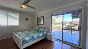 Ein Schlafzimmer mit Bett, Nachttisch, Deckenventilator, Fenster mit Jalousien und Blick auf eine Terrasse und ein Gebäude.