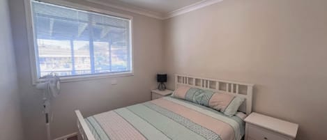 A bedroom with a bed, bedside tables, a standing fan, and a window with blinds.
