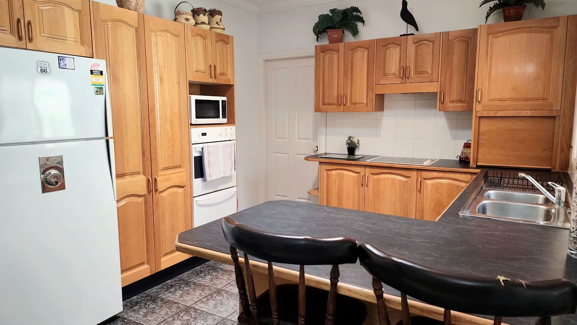 Une cuisine avec des meubles de rangement en bois, un réfrigérateur, un four à micro-ondes, un four, un évier et deux tabourets de bar.