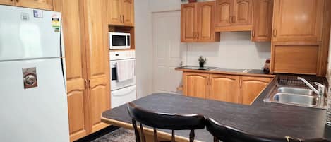 Une cuisine avec des meubles de rangement en bois, un réfrigérateur, un four à micro-ondes, un four, un évier et deux tabourets de bar.