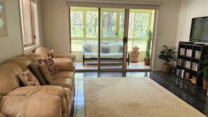 Un salón con un sofá de cuero color beige, una alfombra grande, una estantería y un ventanal corredero que da a un balcón con vistas a árboles.