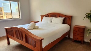 A wooden bed with white linens, a nightstand, and a dresser in a room with a window and a plant.