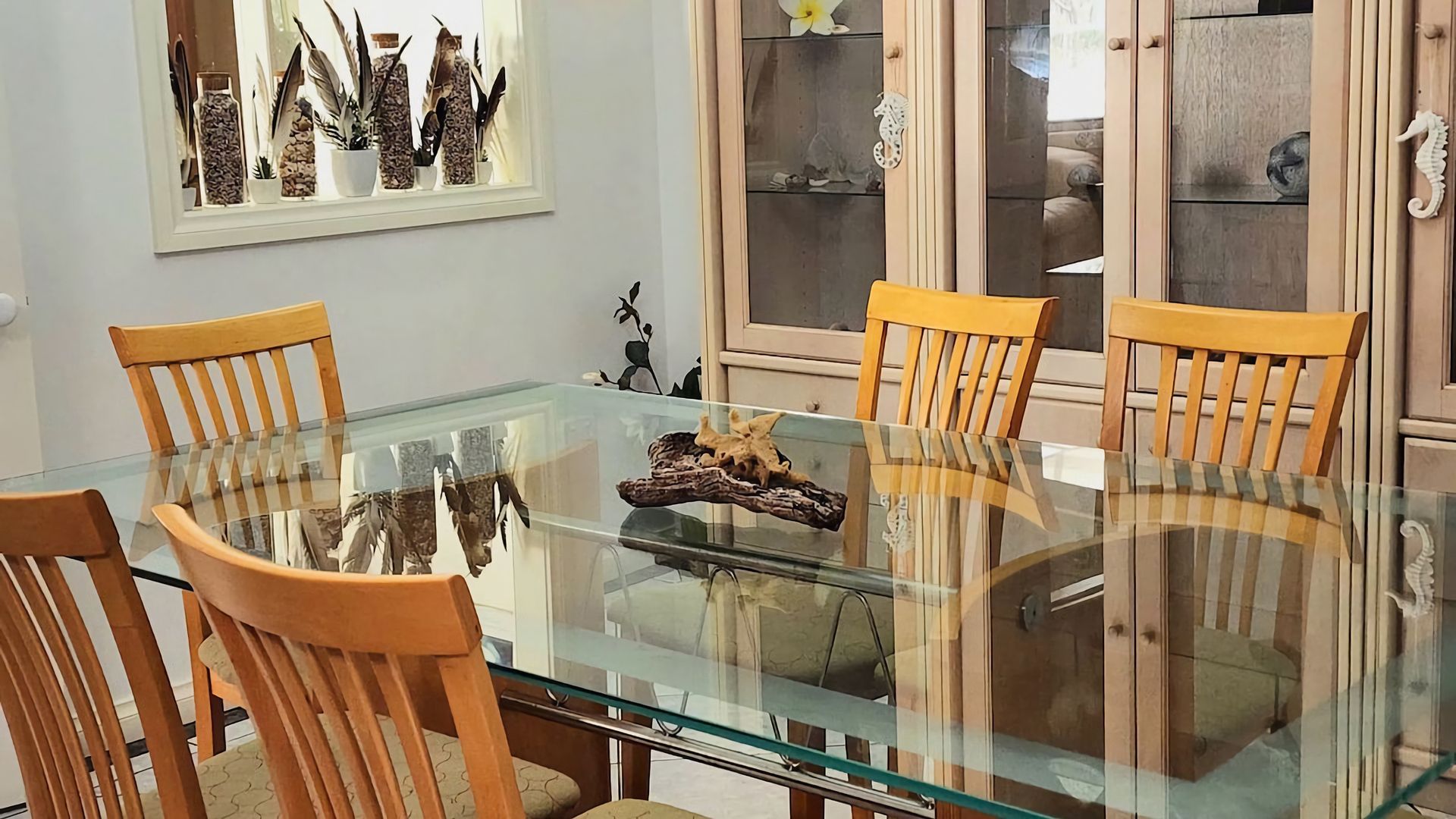 A dining area with a glass table surrounded by wooden chairs, a decorative piece on the table, and a cabinet with glass doors in the background.