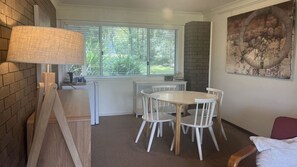 A room with a white table and chairs, a window with blinds, a brick wall, and a painting on the wall. - The Sea Shack at Berrara (Berrara)