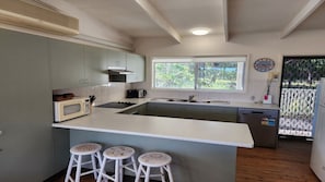 A kitchen with a central island, white stools, a microwave, a stove, and a window with a view of greenery. - The Sea Shack at Berrara (Berrara)