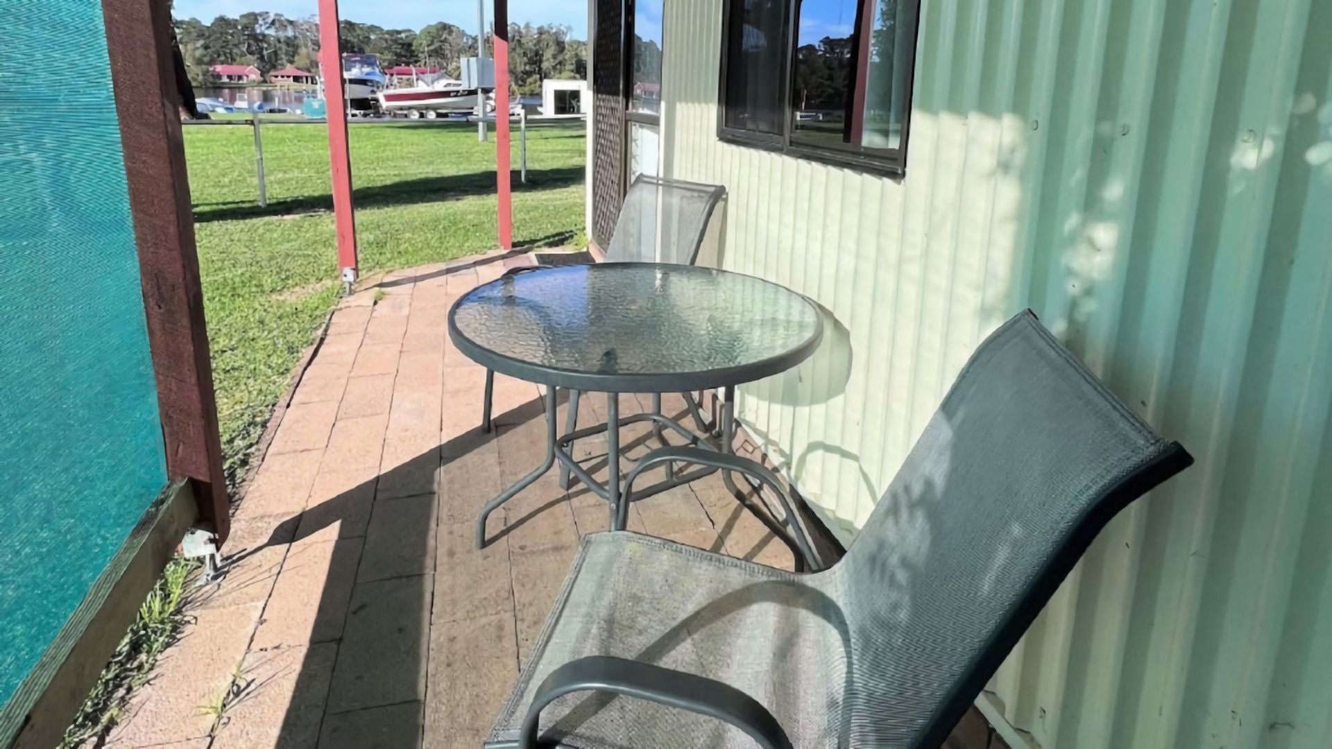 A patio with a glass table and metal chairs, a small covered area, and a view of a grassy area with boats.