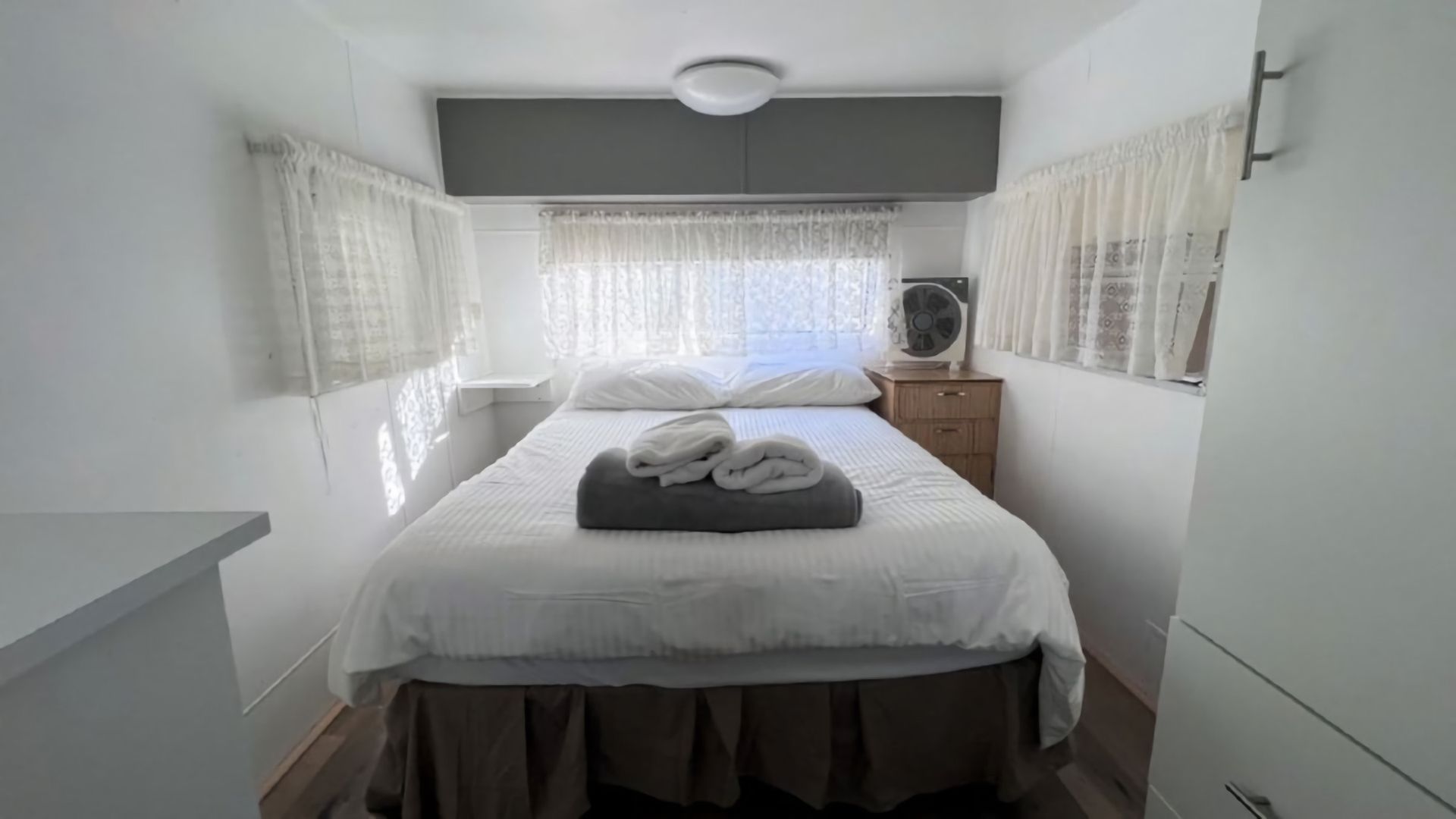 A bedroom with a bed, a bedside table, a fan, and curtains.