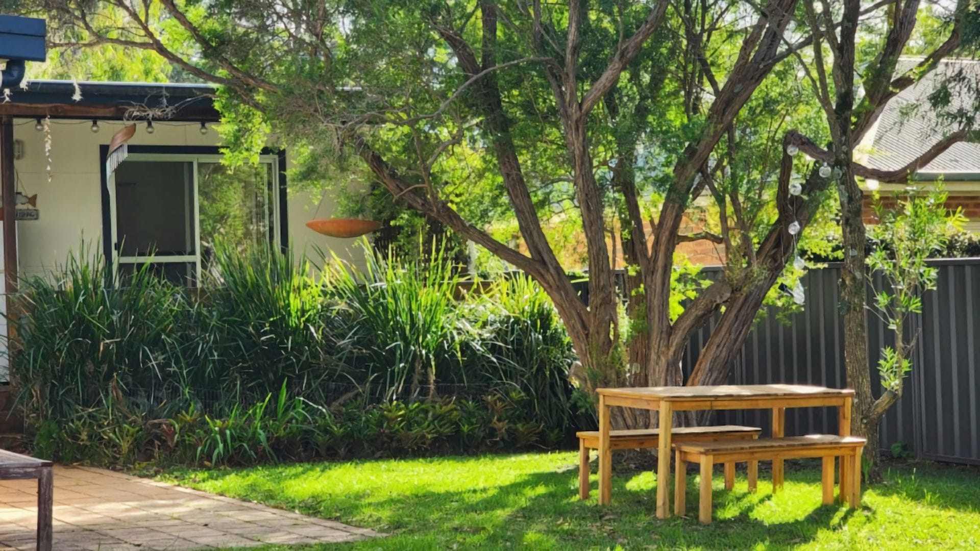 A wooden bench and table set in a garden with a tree and a fence in the background.