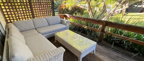 Een overdekt terras met een bank en tafel, omgeven door een houten terras en beplanting.