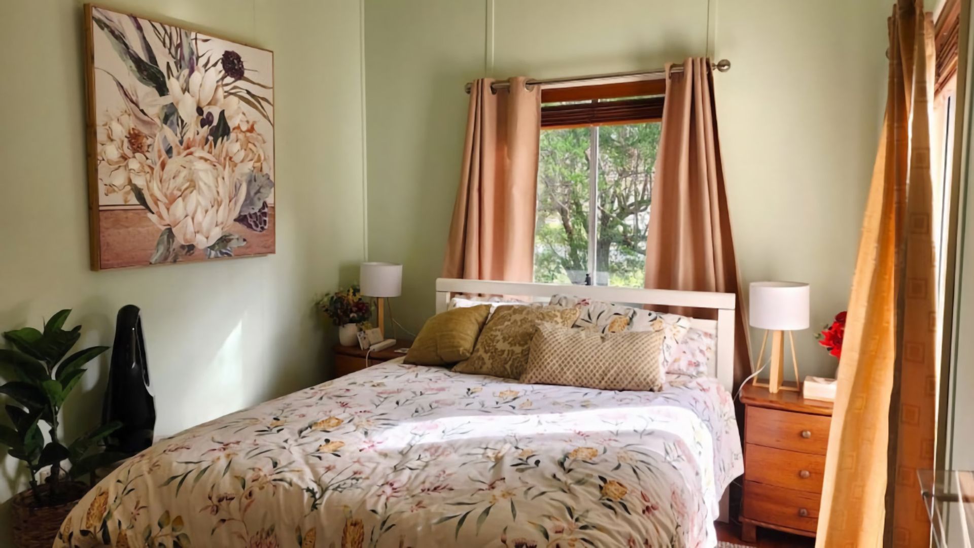 Ein Schlafzimmer mit einem Bett, einem Nachttisch, einer Lampe, einem Blumentopf und einem Fenster mit Vorhängen.