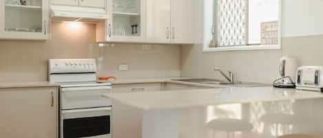 Una cocina con estufa, horno y pileta. Hay alacenas sobre la encimera y una ventana con un panel decorativo.