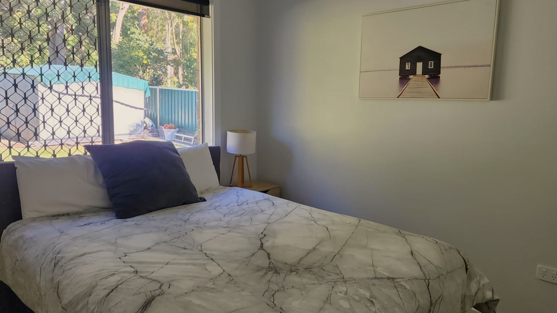 A bedroom with a bed, a nightstand, a lamp, and a window with a view of a blue fence and trees.