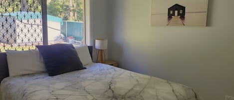 A bedroom with a bed, a nightstand, a lamp, and a window with a view of a blue fence and trees.