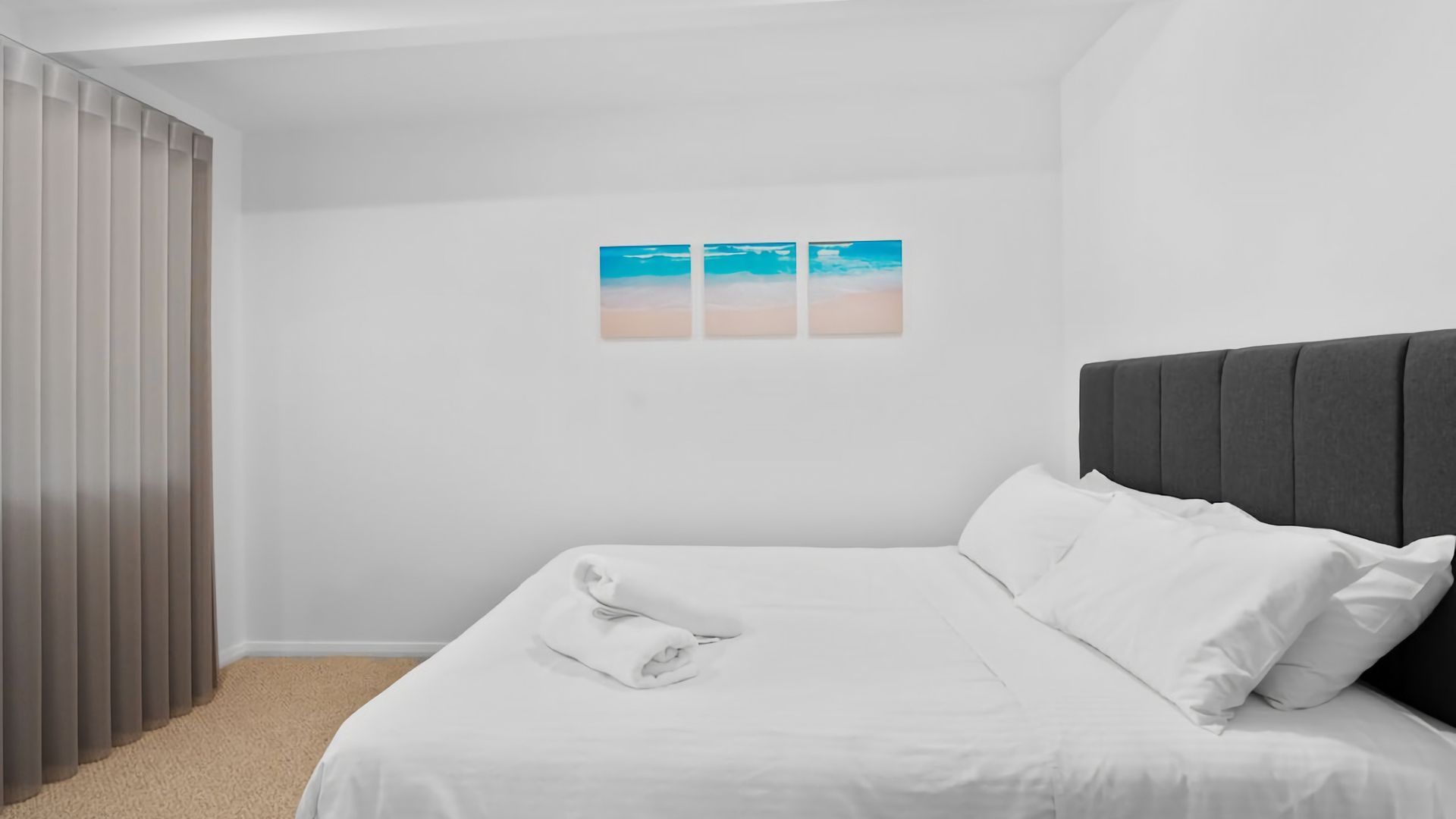 A bedroom with a bed, white bedding, a grey headboard, and a wall with three framed pictures.