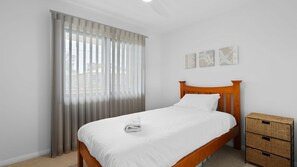 Cama bem arrumada com cabeceira de madeira, um conjunto de cortinas e um pequeno aparador de vime.