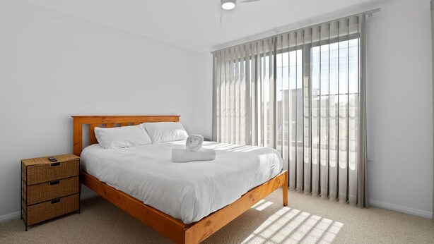 A neatly made bed with white linens and a folded towel, a wooden headboard, a nightstand with drawers, and a large window with vertical blinds. - Sun N Sea in Berrara (Berrara)