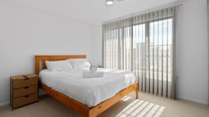 A neatly made bed with white linens and a folded towel, a wooden headboard, a nightstand with drawers, and a large window with vertical blinds.