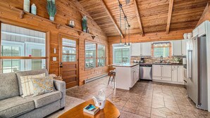 A spacious living area with a wooden ceiling, a sofa with patterned cushions, a coffee table with a book, and a kitchen with white cabinets and a refrigerator.