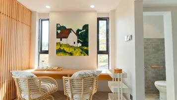 Een moderne kamer met een houten bureau, twee rieten stoelen, een schilderij van een huis, en een badkamer met toilet en douche.