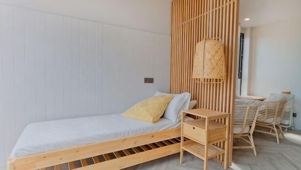 A single bed with a wooden frame, a bedside table, and a chair in a room with white walls and a wooden slat partition. - Talula Hill Farmstay Kluang (Kluang)