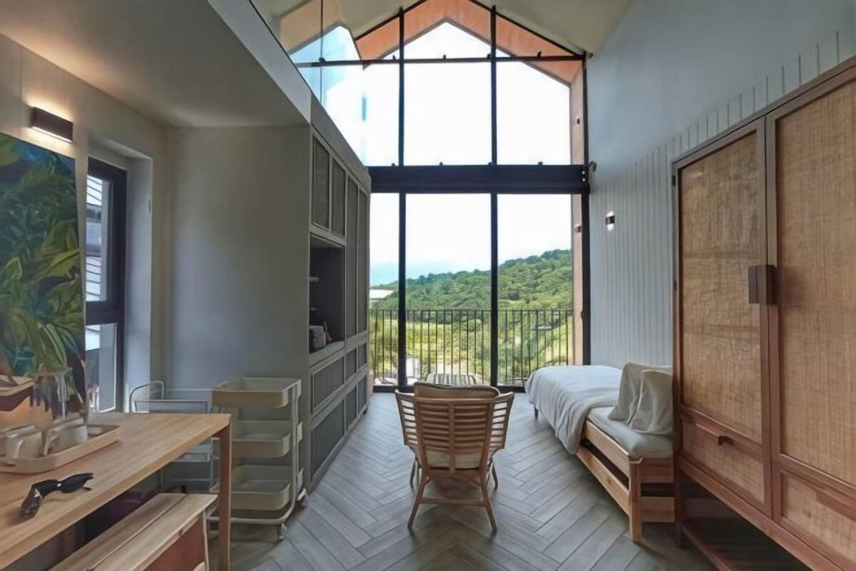 Une pièce avec une grande fenêtre, un lit, une chaise en bois, un bureau et une armoire.
