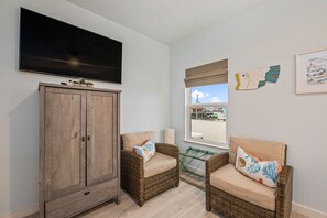 Una sala de estar con un televisor de pantalla plana grande, dos sofás de mimbre con cojines, un aparador de madera y un ventanal con vistas al exterior.
