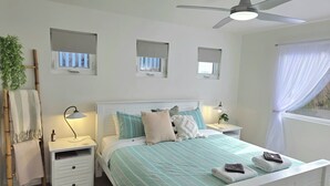 Une chambre à coucher comprenant un lit, des tables de chevet, un ventilateur de plafond et des fenêtres équipées de stores.